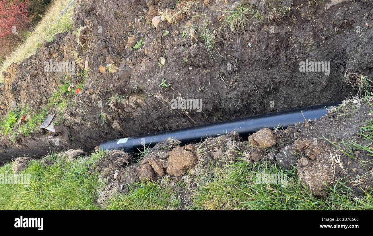 Aushubstelle mit neu installiertem schwarzem Rohr in Graben mit freiliegendem Boden und grünem Gras. Stockfoto