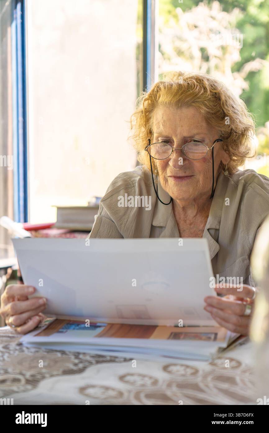 Seniorin mit Brille, überprüft Papiere und Rechnungen, während sie in einem komfortablen häuslichen Umfeld die persönlichen Finanzen verwaltet, wobei Fokus und Stockfoto