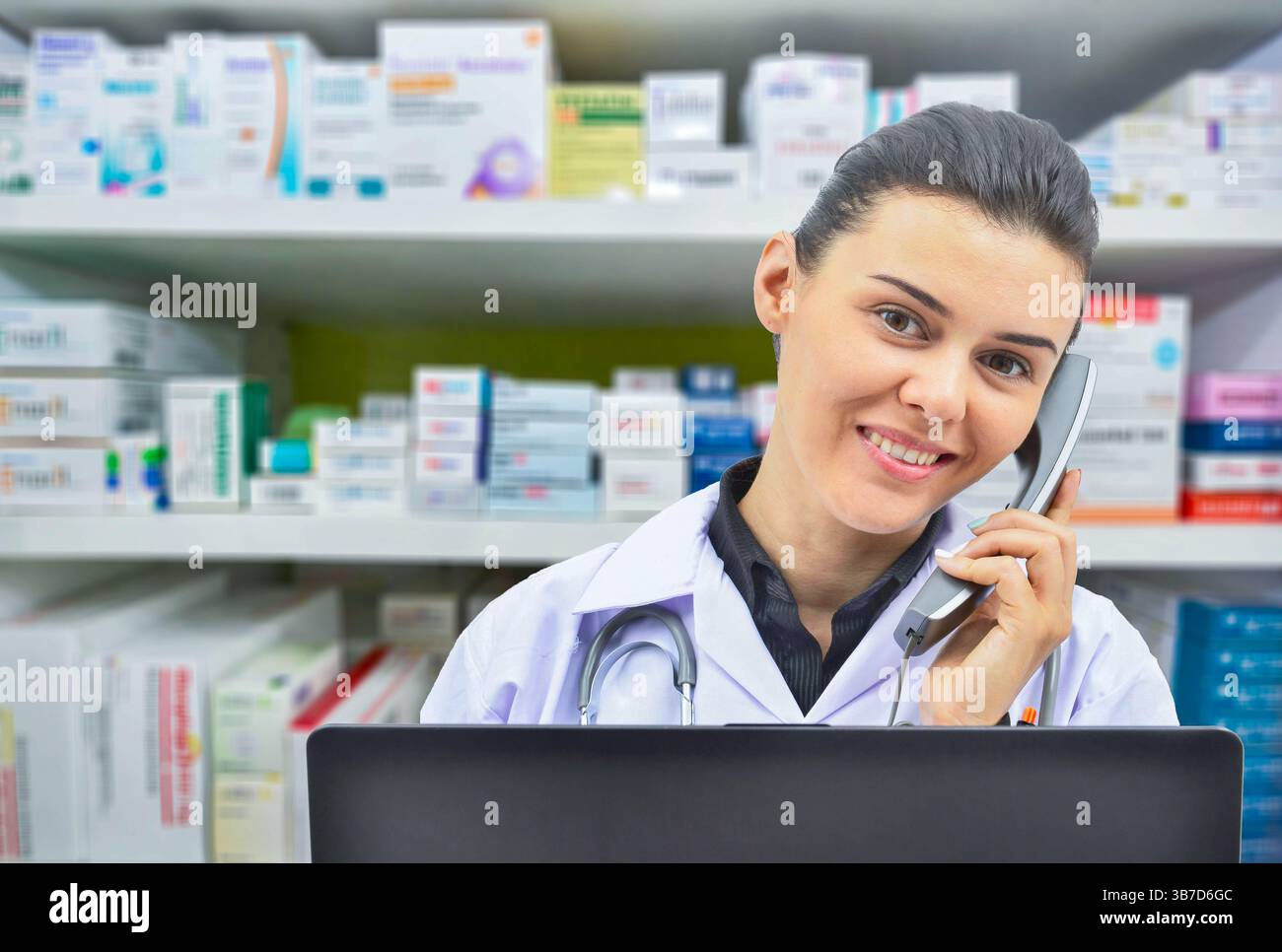 Lächelnde Apothekerin, die mit jemandem am Telefon spricht, während sie einige Bestände im Computer auf Apothekenhintergrund überprüft. Stockfoto