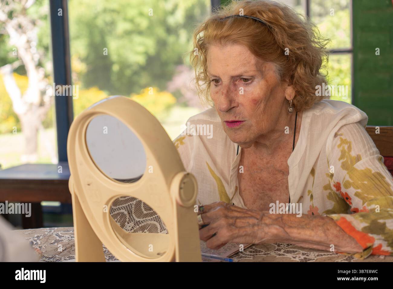 Ältere Frau, die Make-up aufträgt, während sie ihre Reflexion in einem Doppelspiegel auf einem Tisch betrachtet, beleuchtet von hellem natürlichem Licht, das durch einen Gewinn strömt Stockfoto