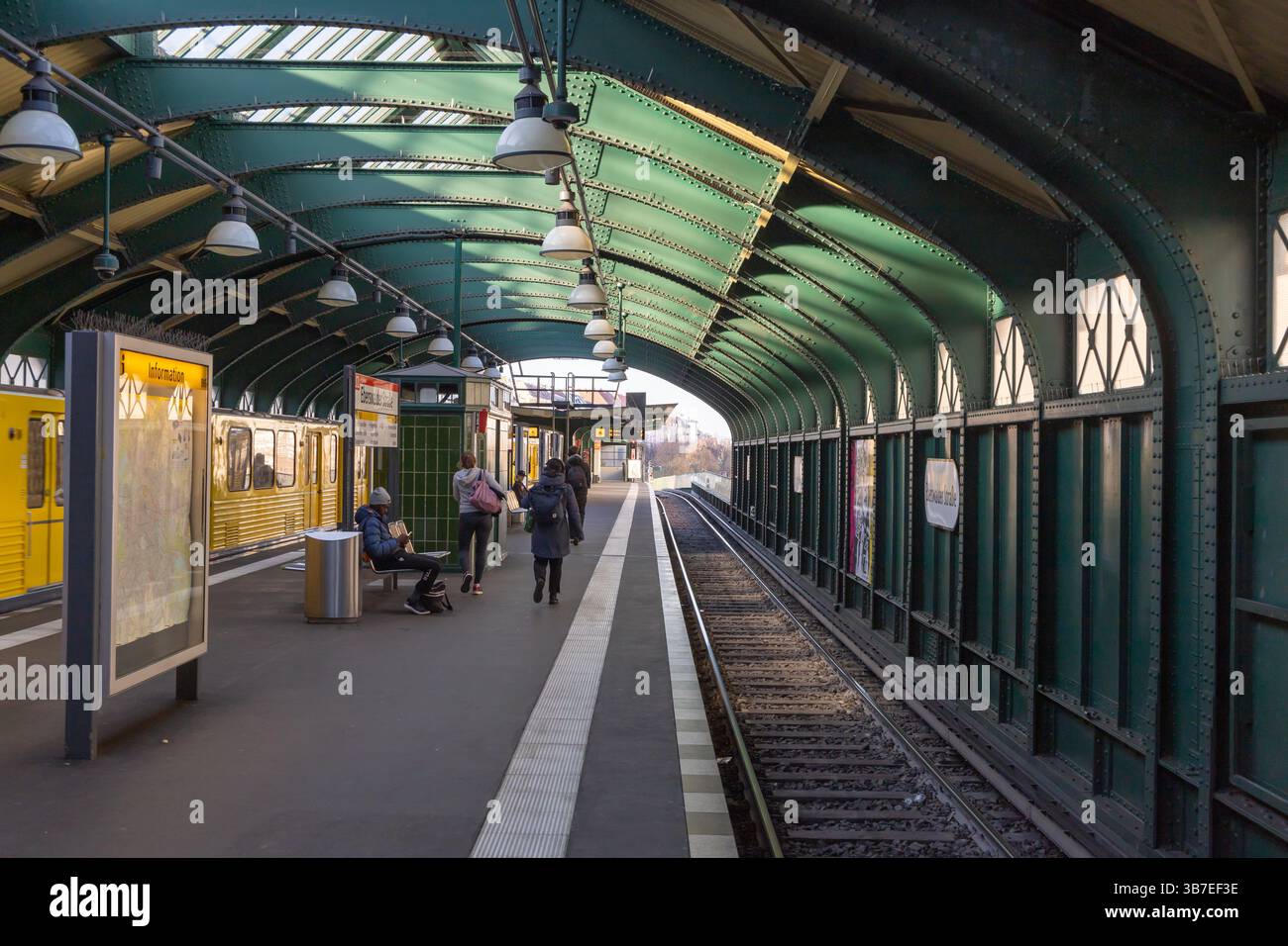 Bahnsteig des U-Bahnhofs Eberswalder Straße in Berlin mit wartenden Fahrgästen und gelbem Zug, der unter dem grünen Metallbogendach ankommt. Ber Stockfoto