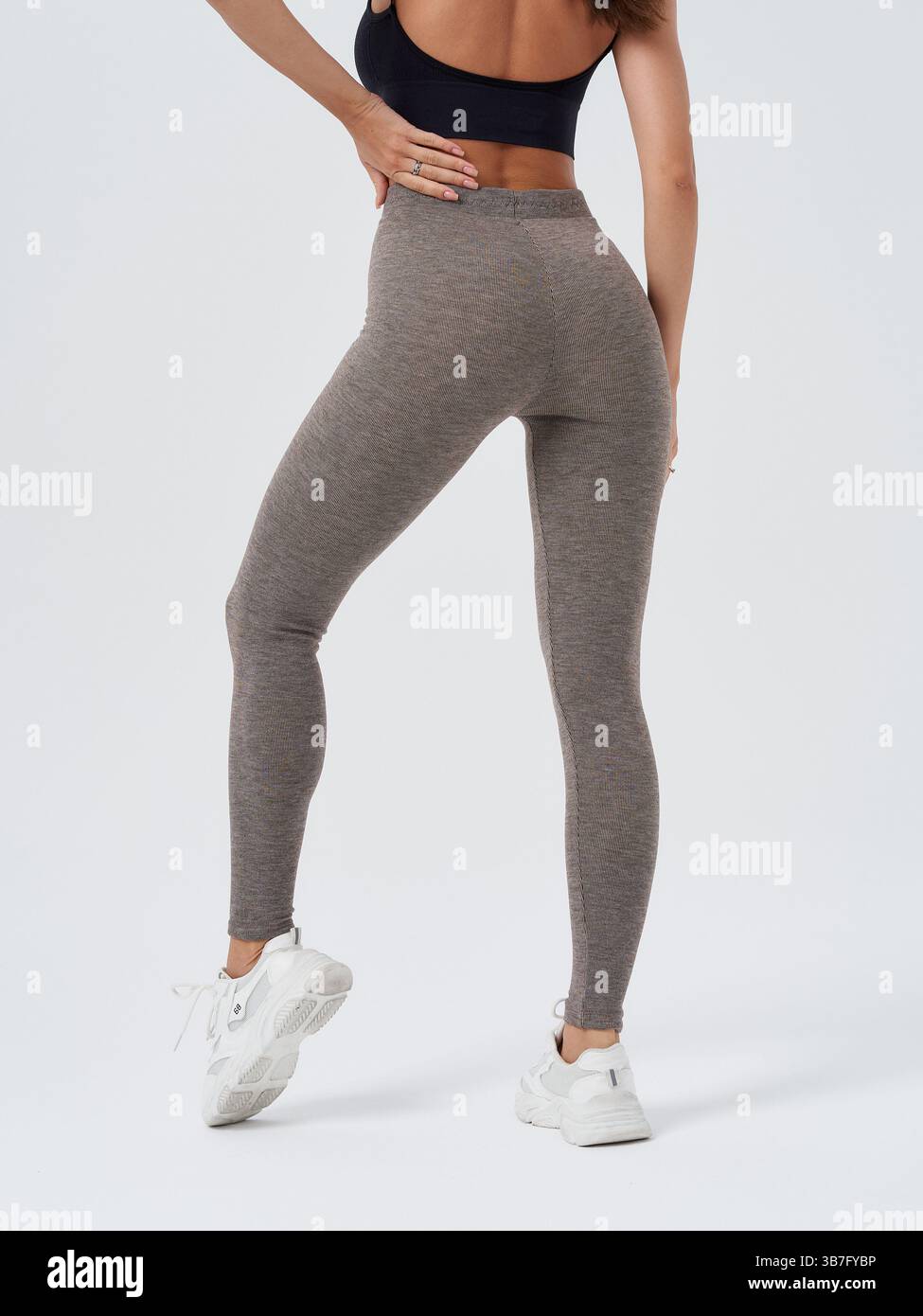 Frau mit grauen Leggings und weißen Turnschuhen, posiert mit Hand auf Hüfte, in einer selbstbewussten Haltung. Schwarzes ärmelloses Crop-Top für sportliche und passgenaue Passform Stockfoto