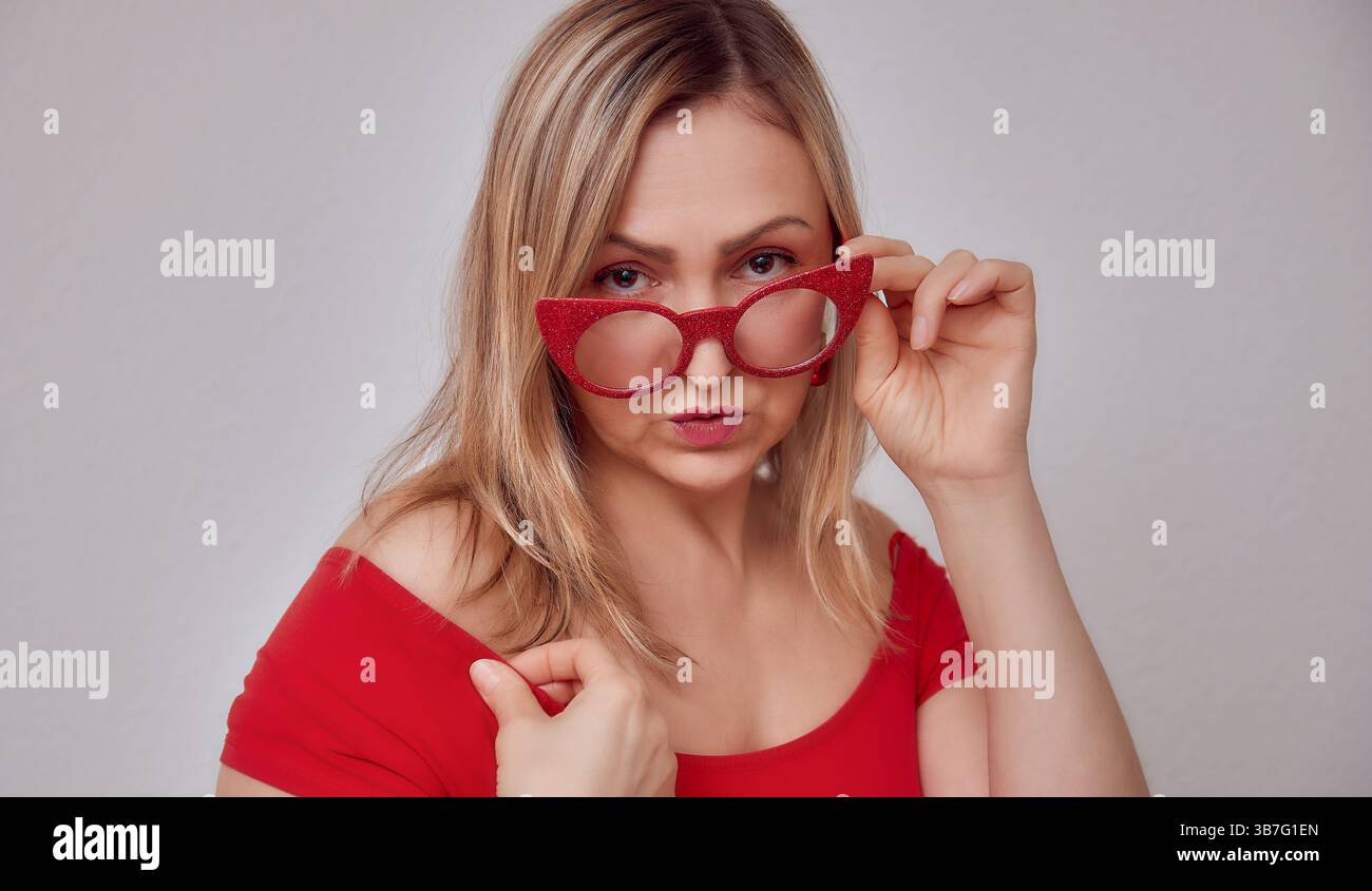 Selbstbewusste blonde Frau in roter Off-Shoulder-Bluse und Brille Back to School Teacher Look, Studio Porträt Stockfoto