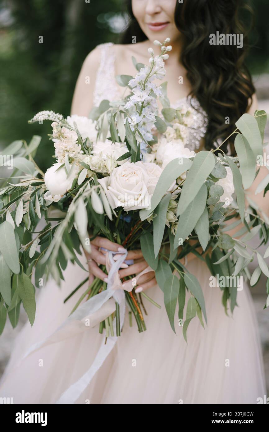 Die Braut steht mit einem Blumenstrauß aus weißen Rosen, Pfingstrosen, Delfinen und Eukalyptuszweigen in der Hand, Nahaufnahme. Stockfoto