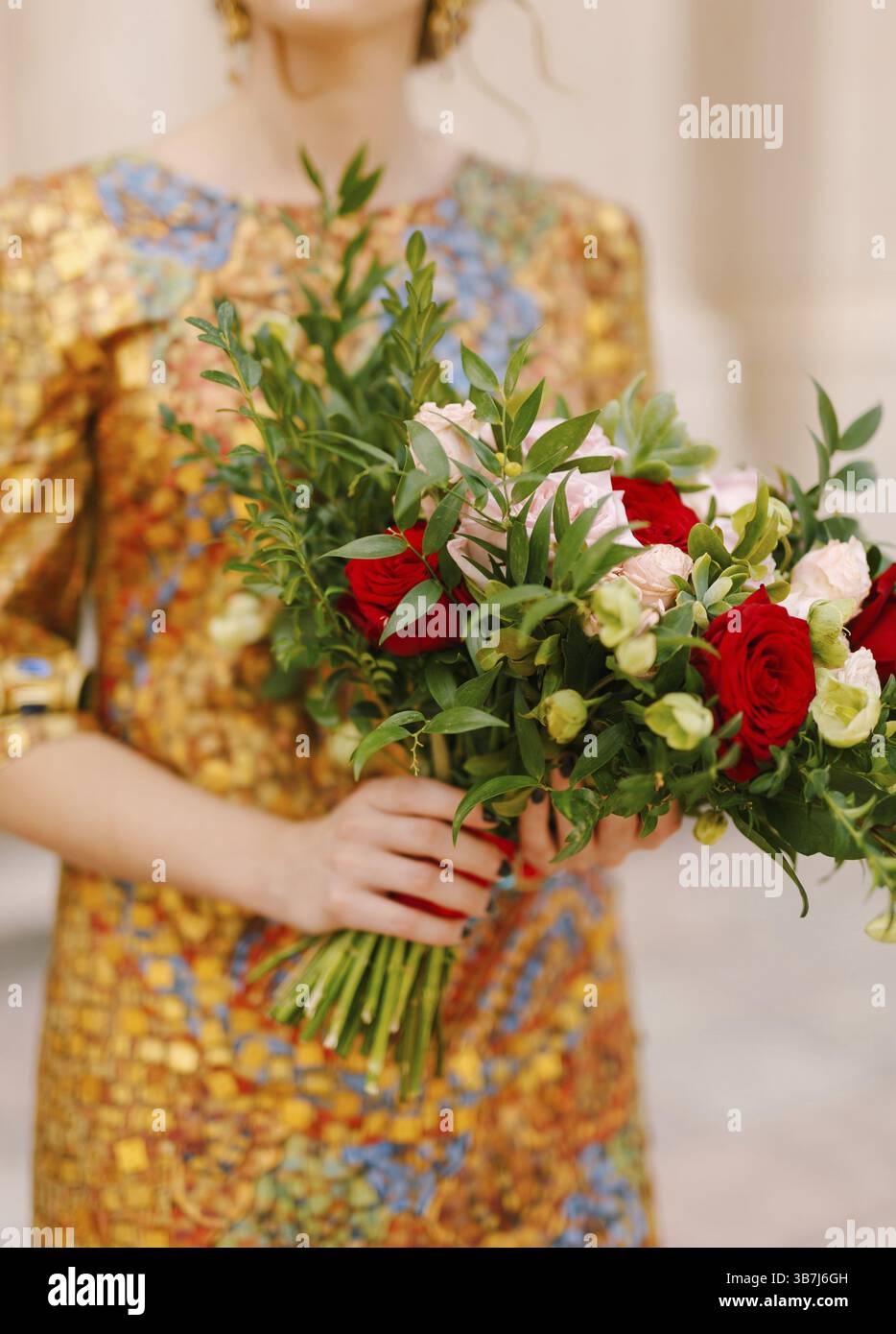 Eine Braut in einem ungewöhnlichen goldenen Kleid hält einen Brautstrauß aus weißen und roten Rosen in der Hand, Nahaufnahme. Stockfoto