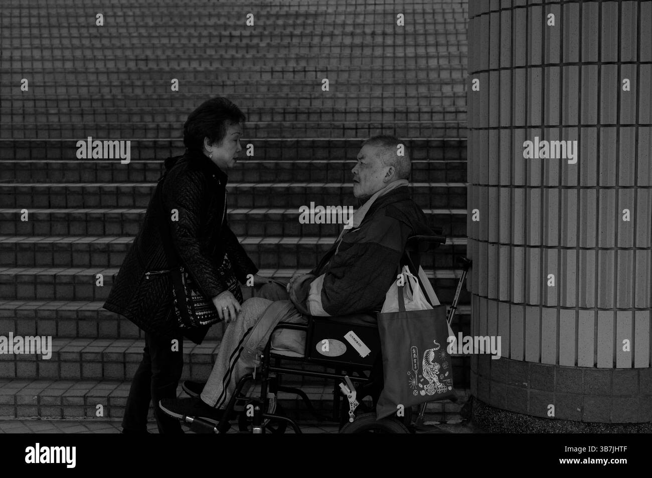 Frau lehnt sich ein, um mit einem Mann im Rollstuhl auf den Stufen des Hong Kong Cultural Centre zärtlich zu sprechen Stockfoto