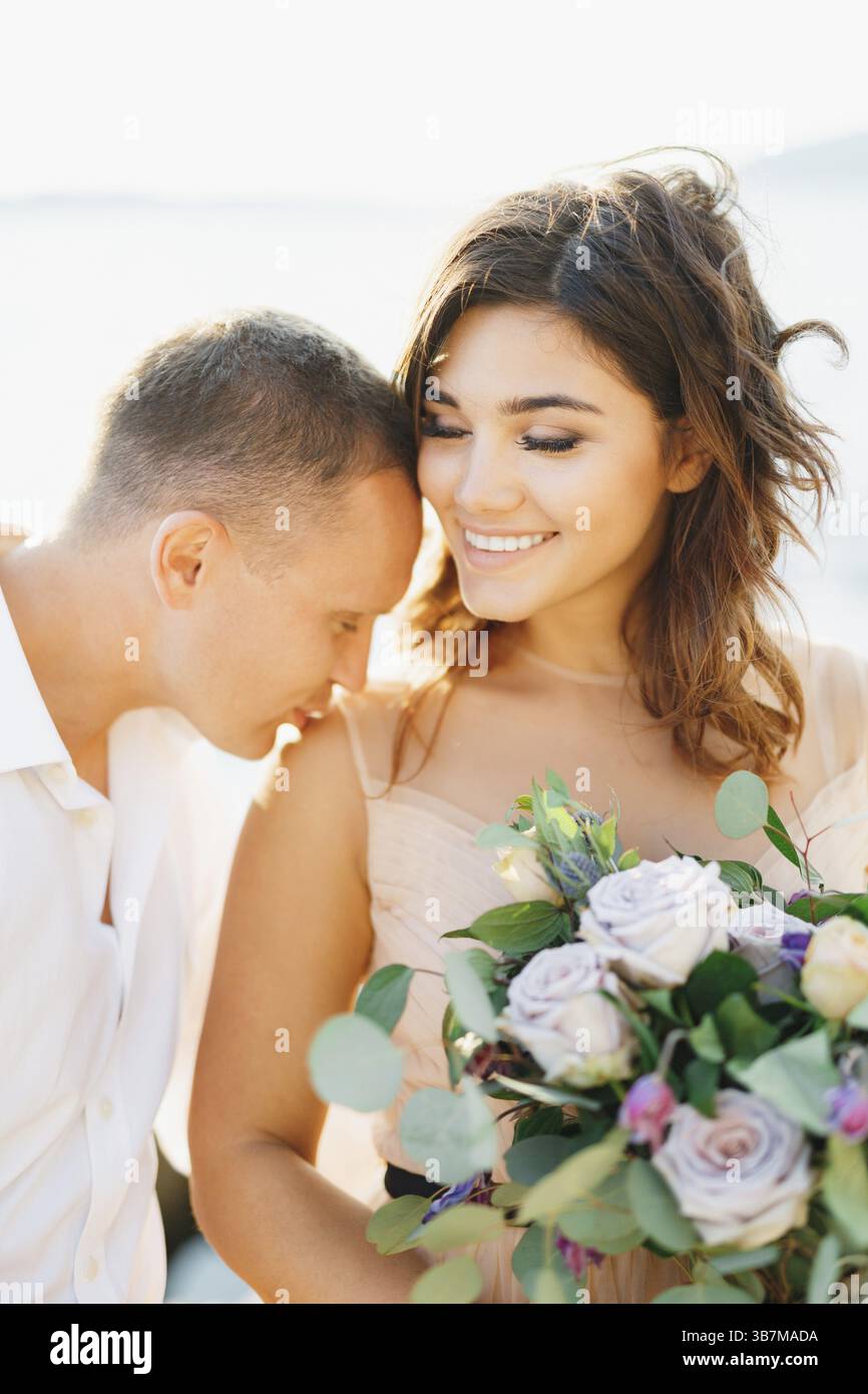 Bräutigam in weißem Hemd küsst auf die Schulter eine lächelnde Braut in einem wunderschönen Pastellkleid mit einem Blumenstrauß in ihren Händen. Stockfoto