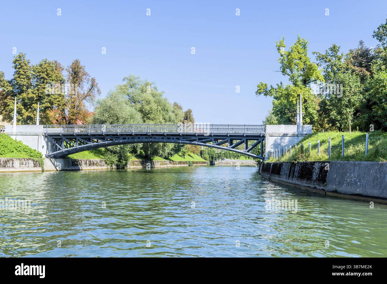 Ljubljana, Slowenien. August 2023. Eine Metallbrücke über den städtischen Wasserweg, gesäumt von üppigem Grün Stockfoto Ljubljana, Slowenien. August 2023. Eine Metallbrücke über den städtischen Wasserweg, gesäumt von üppigem Grün Stockfoto