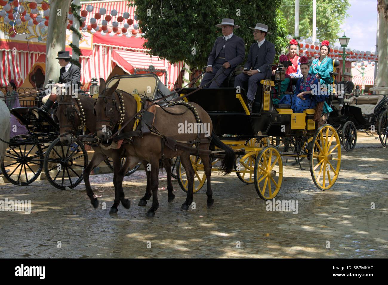 Traditionelle Pferdekutsche auf der Feria de Sevilla 2025 mit Reitern in formeller Kleidung und Frauen in farbenfroher Flamenco-Kleidung, die die festliche Parade genießen. Stockfoto