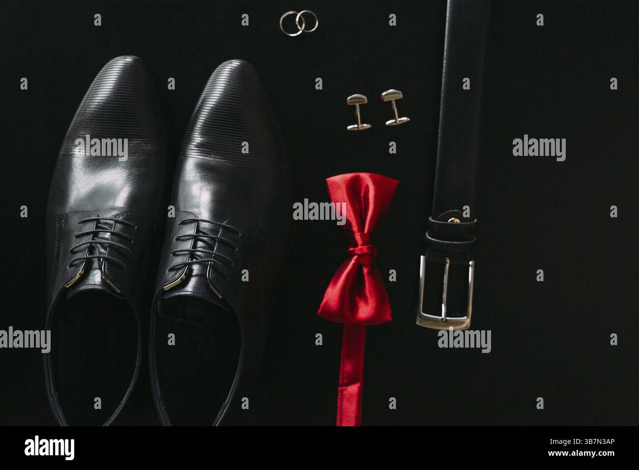 Schwarze Männerschuhe, Manschettenknöpfe, Eheringe, ein schwarzer Gürtel und eine rote Fliege auf schwarzem Hintergrund Stockfoto