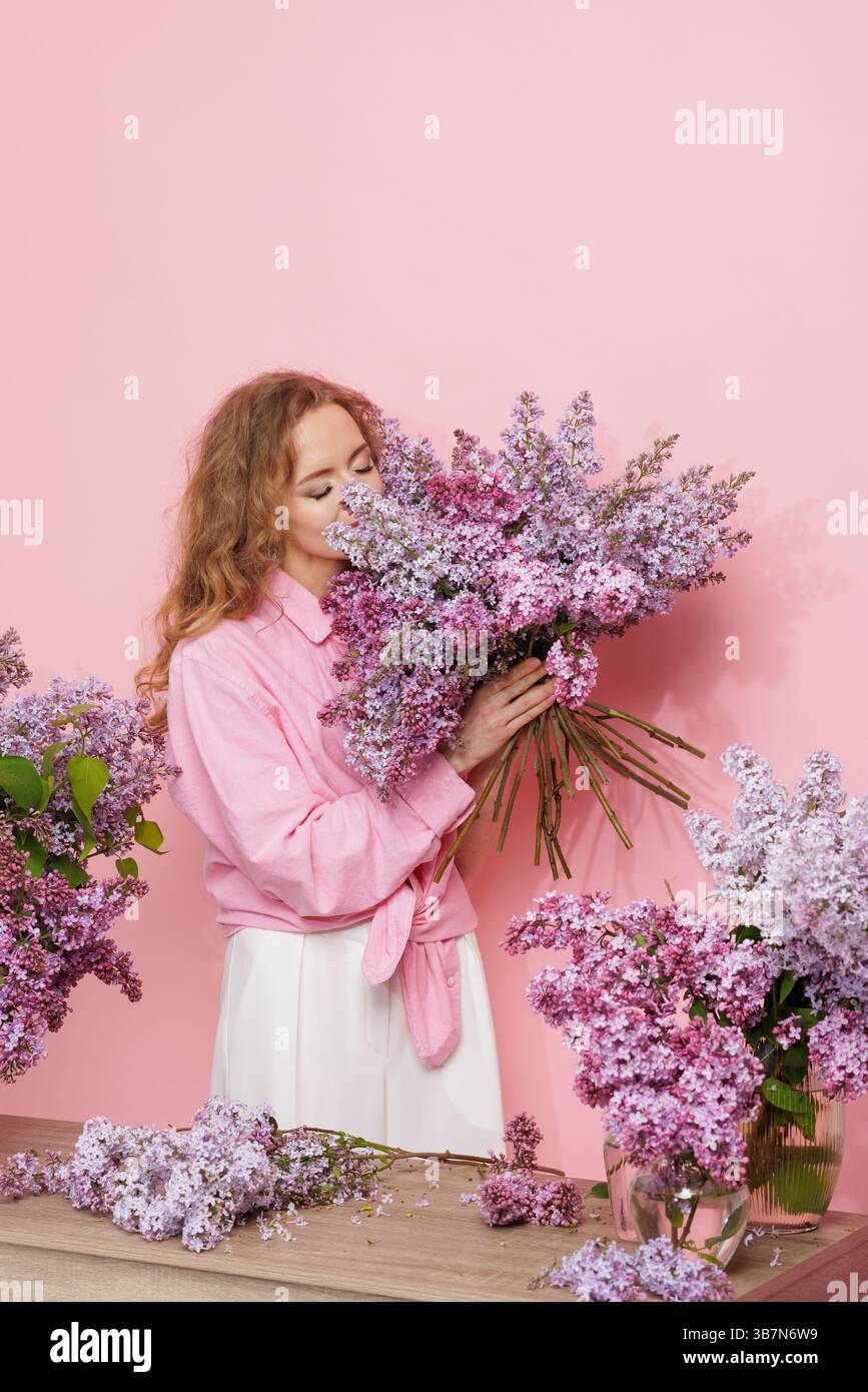 Fröhliche Frau genießt duftenden Fliederstrauß in einem Frühlingsblumenstudio mit weichem pastellrosa Hintergrund. Stockfoto