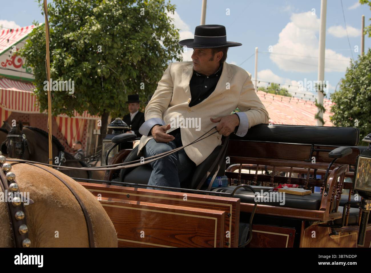 Andalusischer Kutscher in leichter Jacke und Hut, der während der Feria de Sevilla 2025 eine Pferdekutsche fährt. Feria de Sevilla, Spanien Stockfoto