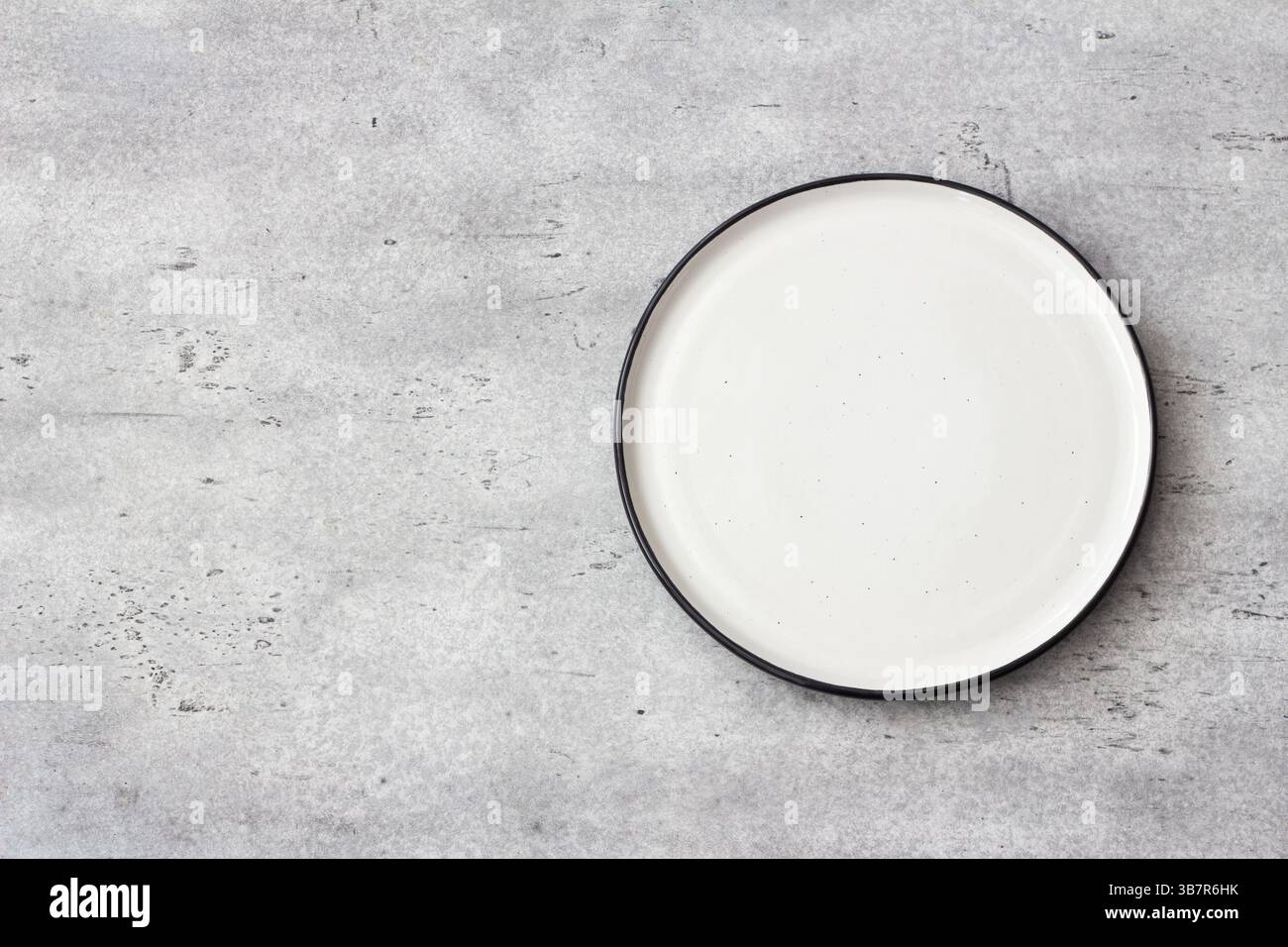 Leere runde weiße Keramikplatte mit schwarzem Rand auf betongrauem texturiertem Hintergrund. Minimalistischer Tisch, Draufsicht. Ideal für Food Mockups und DE Stockfoto