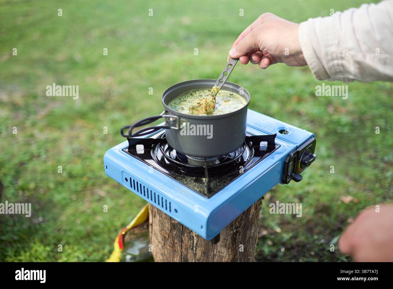 Nicht erkennbare Person, die ein vegetarisches Mittagessen im Freien kocht, Reis mit Gemüse, mit einem Kochtopf auf einem hellblauen tragbaren Gasherd, während sie bei Camping Stockfoto