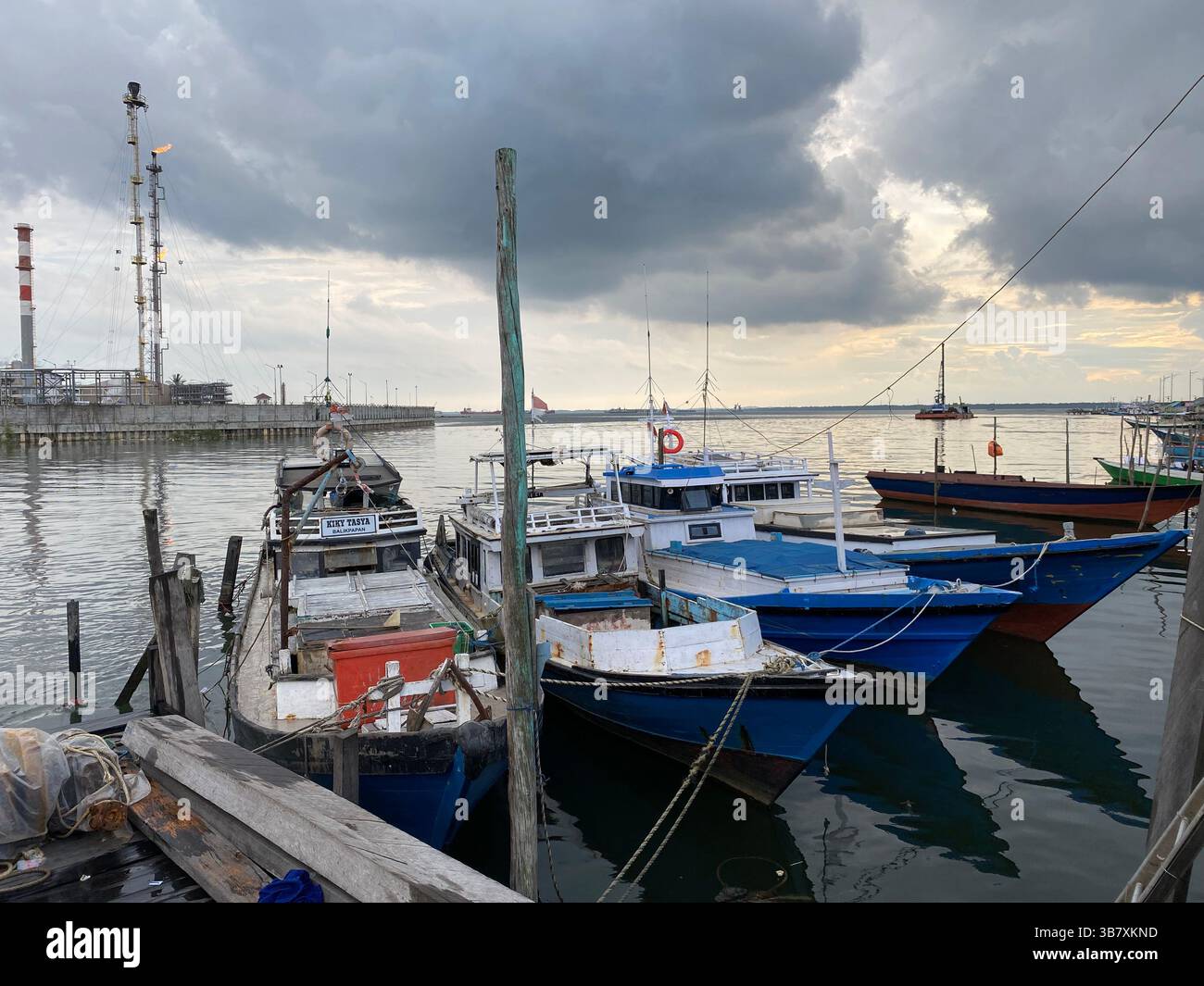 Traditioneller Fischerhafen an der Küste mit Holzbooten, die im Hintergrund von Industrieanlagen mit Schornsteinen und Flare Stacks vertäut sind. - Smartphone-aufgenommenes Stockfoto