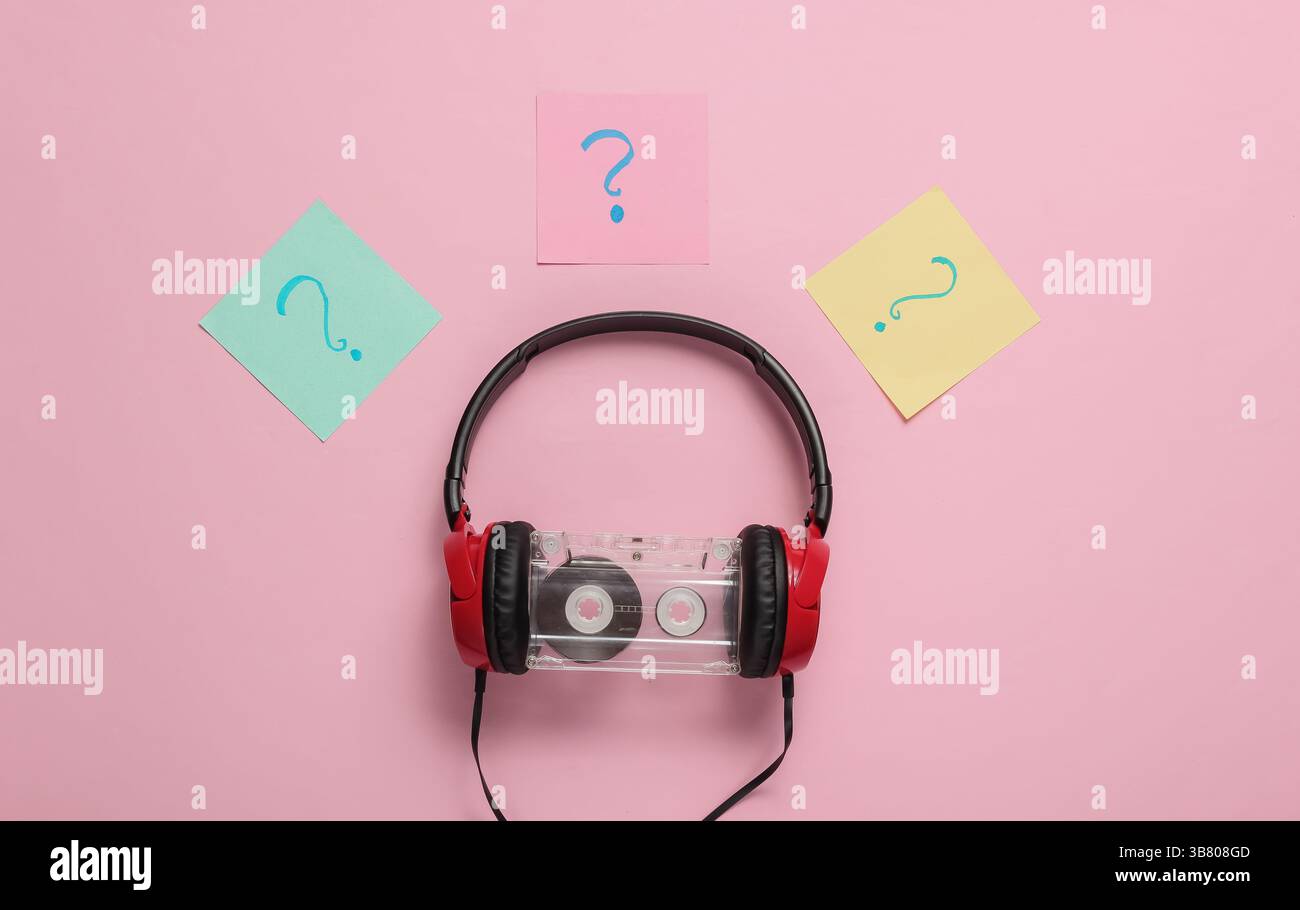 Kabelgebundene Kopfhörer mit Audiokassette und Fragezeichen auf Notizblättern auf rosa pastellfarbenem Hintergrund. Retro-Style, DJ. Blick von oben, minimalistischer Mus Stockfoto