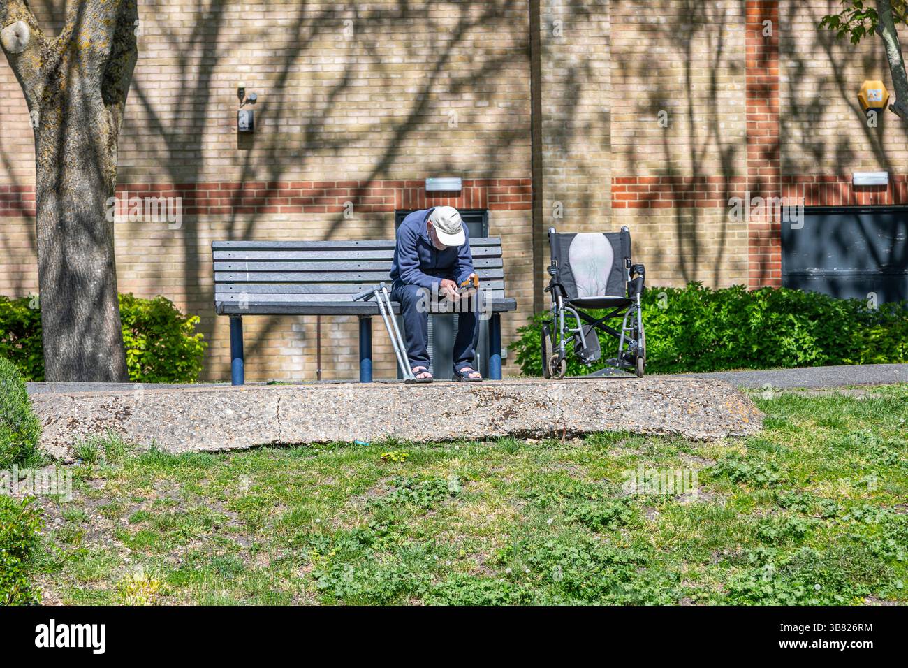 Ein einsamer alter Mann mit Rollstuhl und Krücken, der auf einer Bank in der Sonne sitzt. Stockfoto