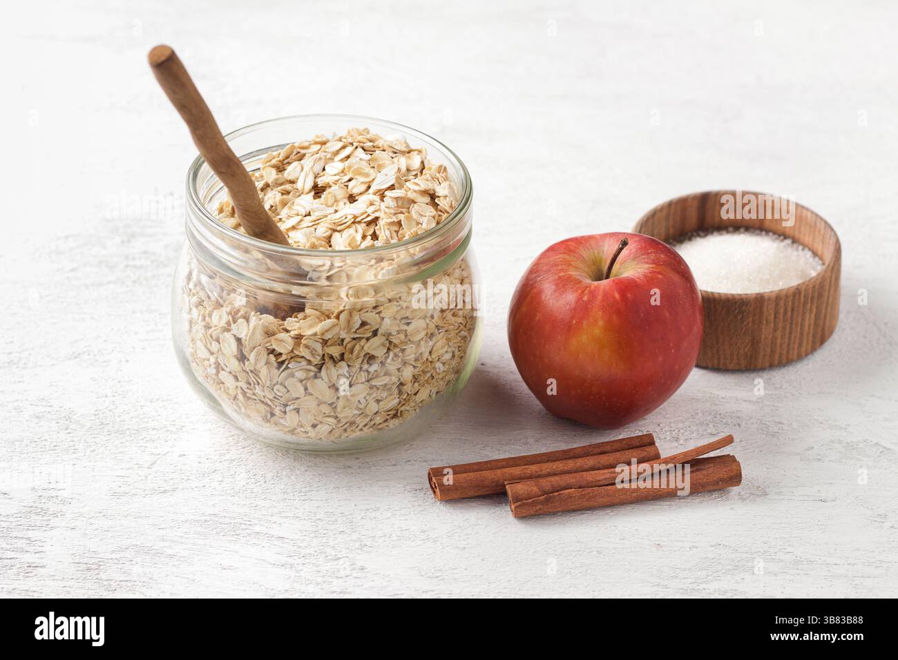 Rohe Haferflocken in einem Glasgefäß mit Zimtstangen, Äpfeln und Zucker auf einem hellgrauen strukturierten Hintergrund. Stockfoto