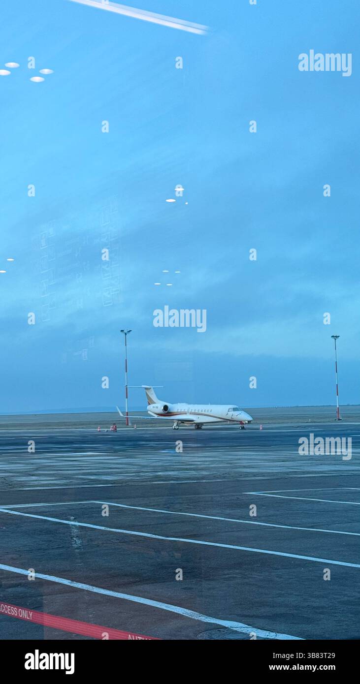 Privatjet parkt auf dem Asphalt eines Flughafens unter klarem blauem Himmel, der vor der Abfahrt am frühen Morgen durch ein Terminal mit Fenstern betrachtet wird. Stockfoto