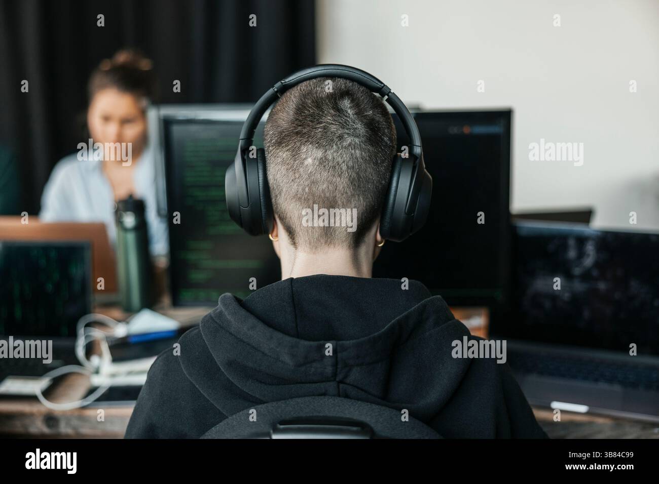Rückansicht eines nicht binären IT-Profis, der während der Arbeit im Büro kabellose Kopfhörercodierung auf einem Desktop-PC trägt Stockfoto