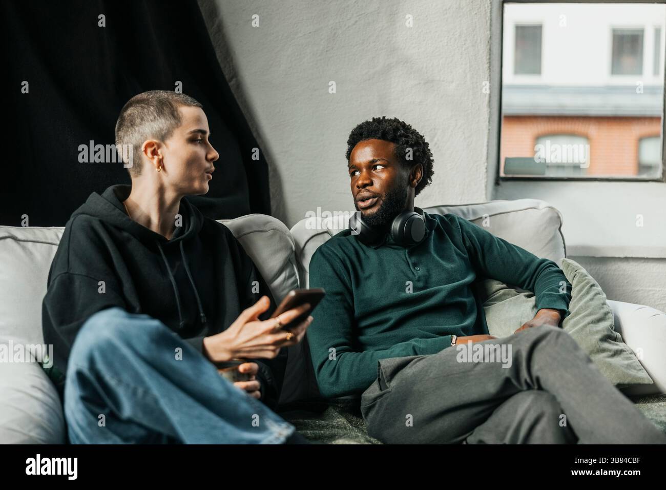 Nicht binäre IT-Profis, die ihr Smartphone halten, während sie mit einem männlichen Kollegen sprechen, während sie im Büro auf dem Sofa sitzen Stockfoto