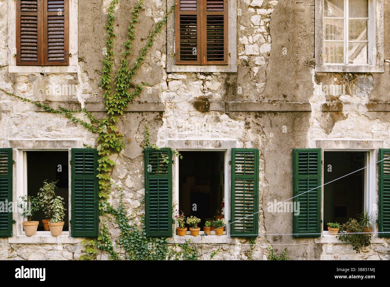 Ein altes Wohngebäude in Rissen mit Fenstern und Fensterläden und Blumen auf den Fensterbänken. Stockfoto