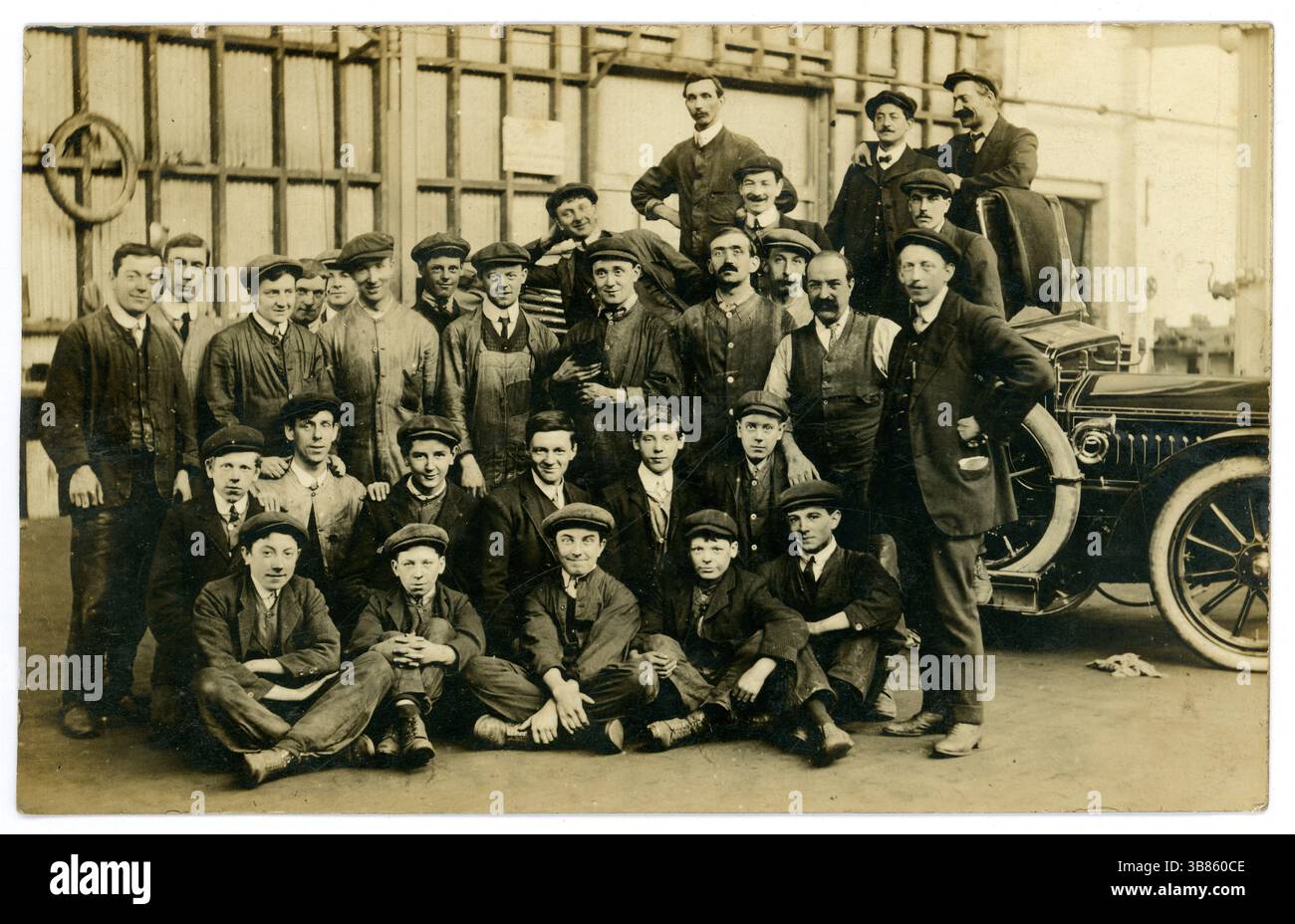 Ursprünglich Anfang der 1900er Jahre, Postkarte aus der Edwardianischen Ära von Werkstattarbeitern, Mechanikern, frühen Motorwagenbauern, in einer großen Werkstatt, in einer Garage, jungen Lehrlingen, Mann mit Katze, viele freche Charaktere. Ein Auto ist sichtbar, möglicherweise ein beliebtes Auto der damaligen Zeit wie der Ford Model T. U.K. um 1910. Stockfoto