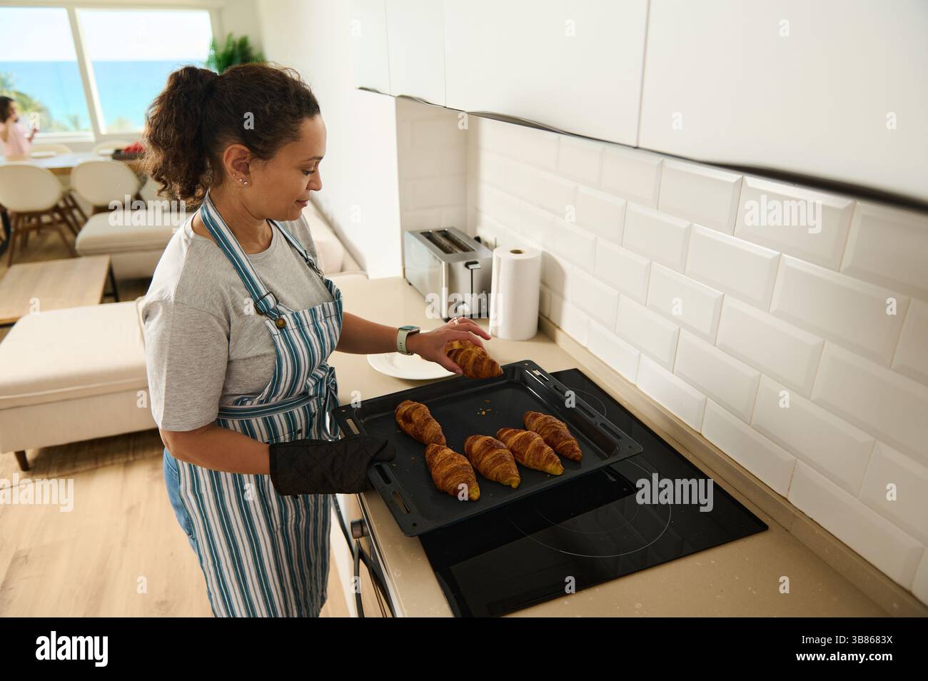 Eine Mutter bereitet in einer gut beleuchteten Küche frisch gebackene Croissants zu, während ihre Familie gemeinsam frühstückt. Der offene Raum und das natürliche Licht schaffen Stockfoto