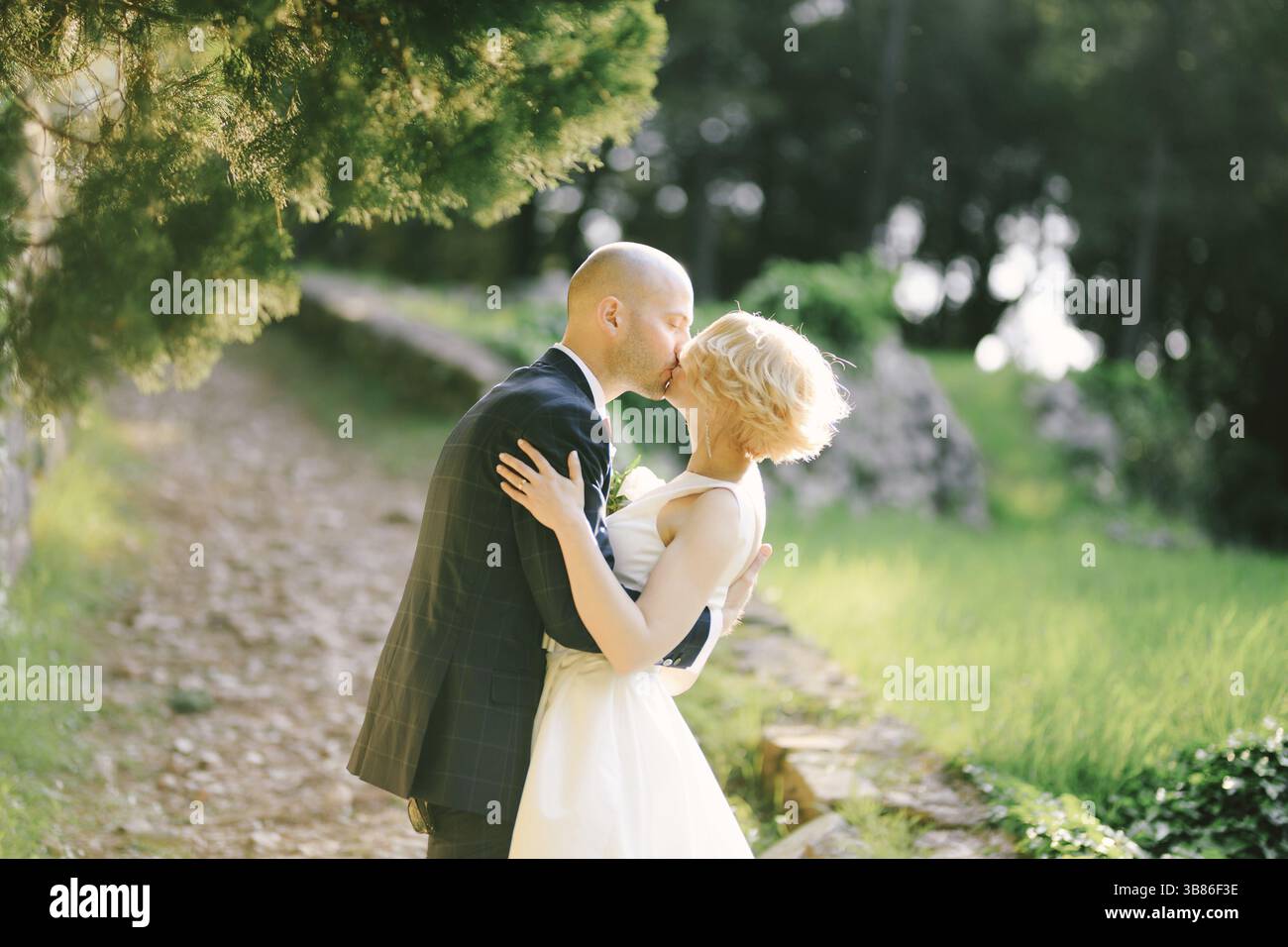 Bräutigam in einem schwarzen Anzug umarmt und küsst Braut in einem weißen Kleid, steht auf einem Pfad im Park vor dem Hintergrund grüner Bäume. Stockfoto