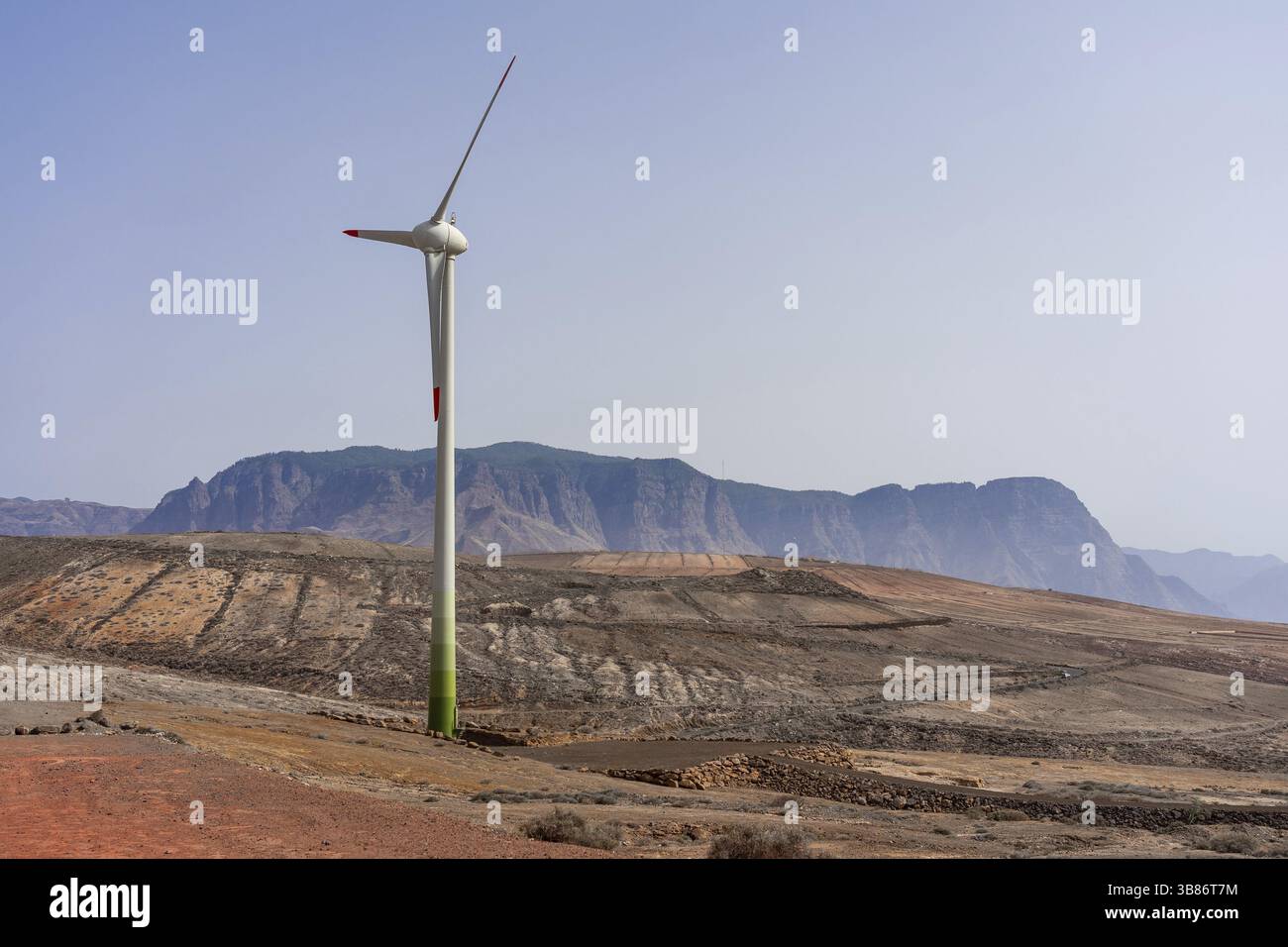 Eine Windkraftanlage, die hoch vor einem Hintergrund aus Bergen steht. Die Landschaft wirkt ruhig, der Windgenerator steht im Fokus und symbolisiert erneuerbares ener Stockfoto