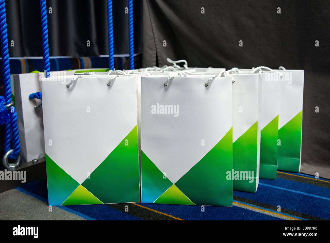 Geschenktüten aus Papier mit mehreren Marken und grünen geometrischen Mustern, die auf einem Teppichboden bei einer Unternehmens- oder Werbeveranstaltung ordentlich angeordnet sind. Stockfoto