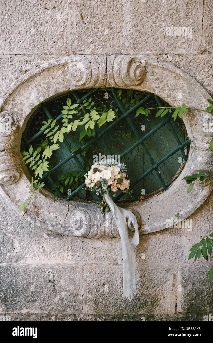 Brautstrauß aus weißen Rosen, Eukalyptuszweigen, sempervivum, scabiosa und blauen Bändern am ovalen Fenster eines alten Hauses. Stockfoto