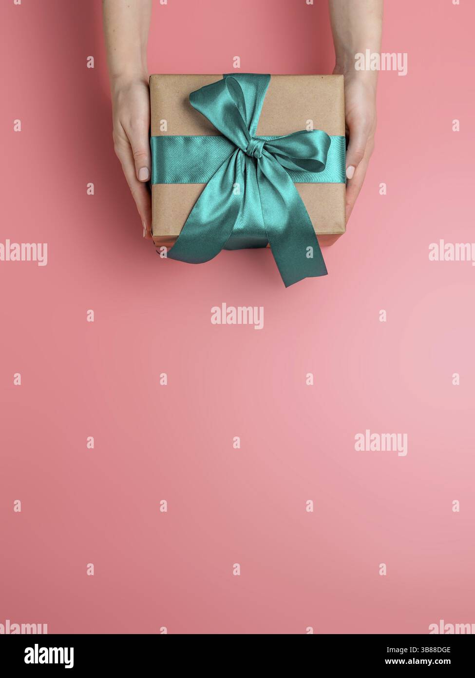 Weibliche Hände halten Geschenkbox auf rosa Hintergrund, kopieren Raum nach unten. Kaukasische Mädchen Hände halten Geschenkbox in Handwerk Verpackung Papier mit grünen Satinband. Stockfoto