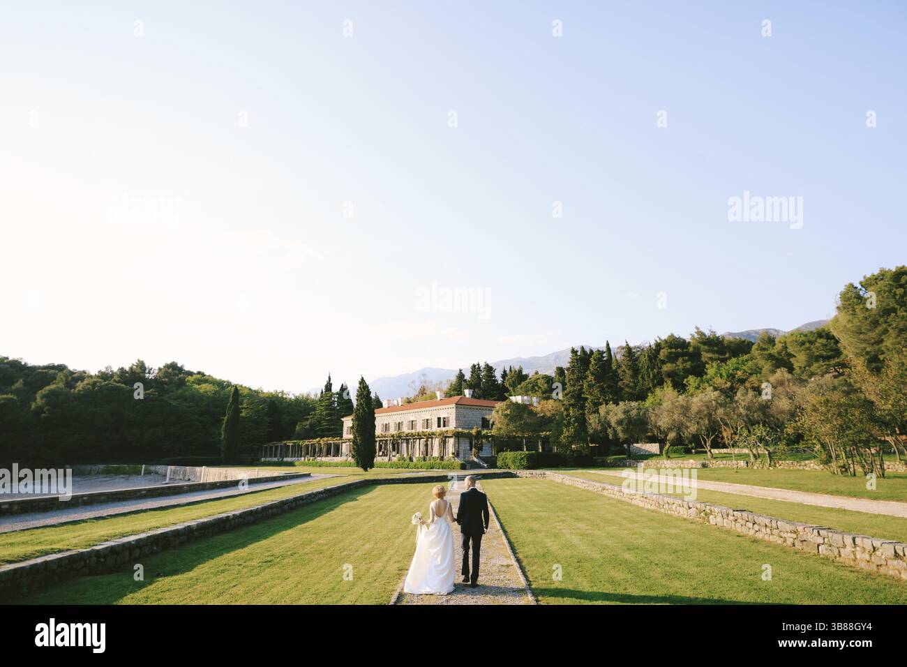 Bräutigam und Braut in einem weißen Kleid mit einem Blumenstrauß gehen in einem schönen Garten vor dem Hintergrund einer alten Villa den Weg entlang. Zurück Stockfoto