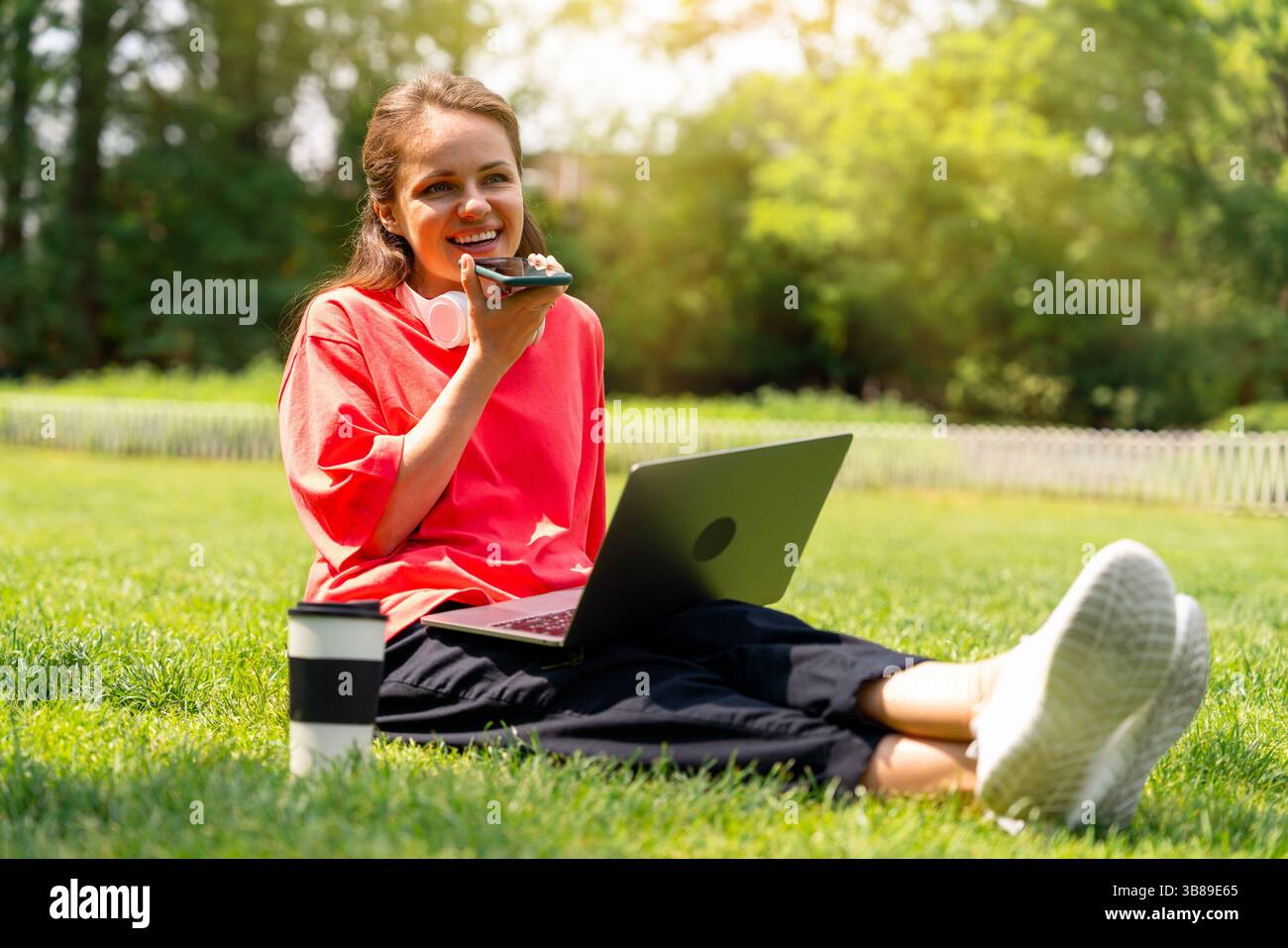 Junge Frau, die Sprachnachrichten aufnimmt, während sie sich mit dem Laptop im Stadtpark entspannt. Digitale Kommunikation. Stockfoto
