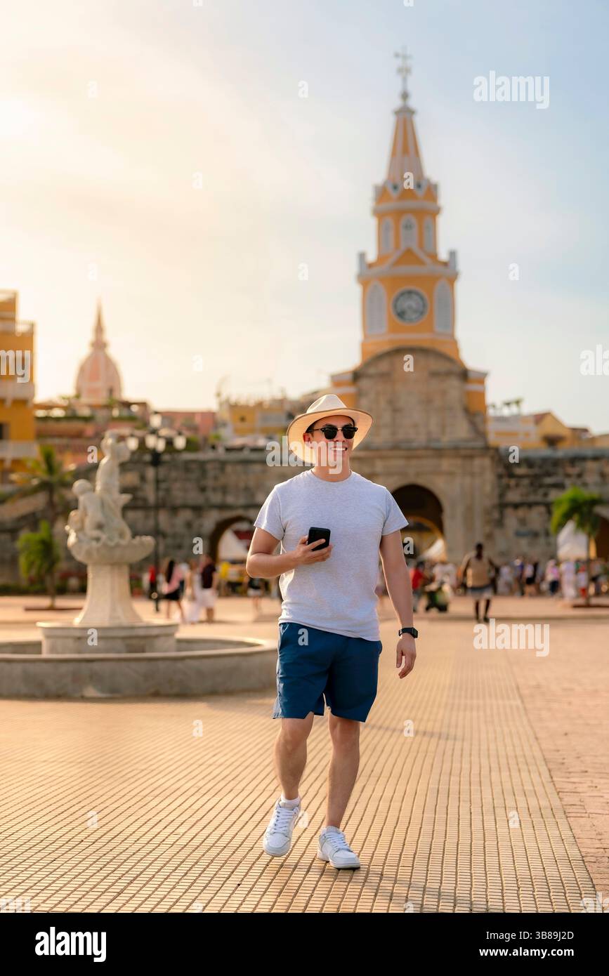 Fröhlicher junger Lateinreisender wandert selbstbewusst durch Cartagena, Kolumbien, trägt eine Sonnenbrille, einen Strohhut und ein lässiges blaues Outfit mit sportlichem Charakter Stockfoto