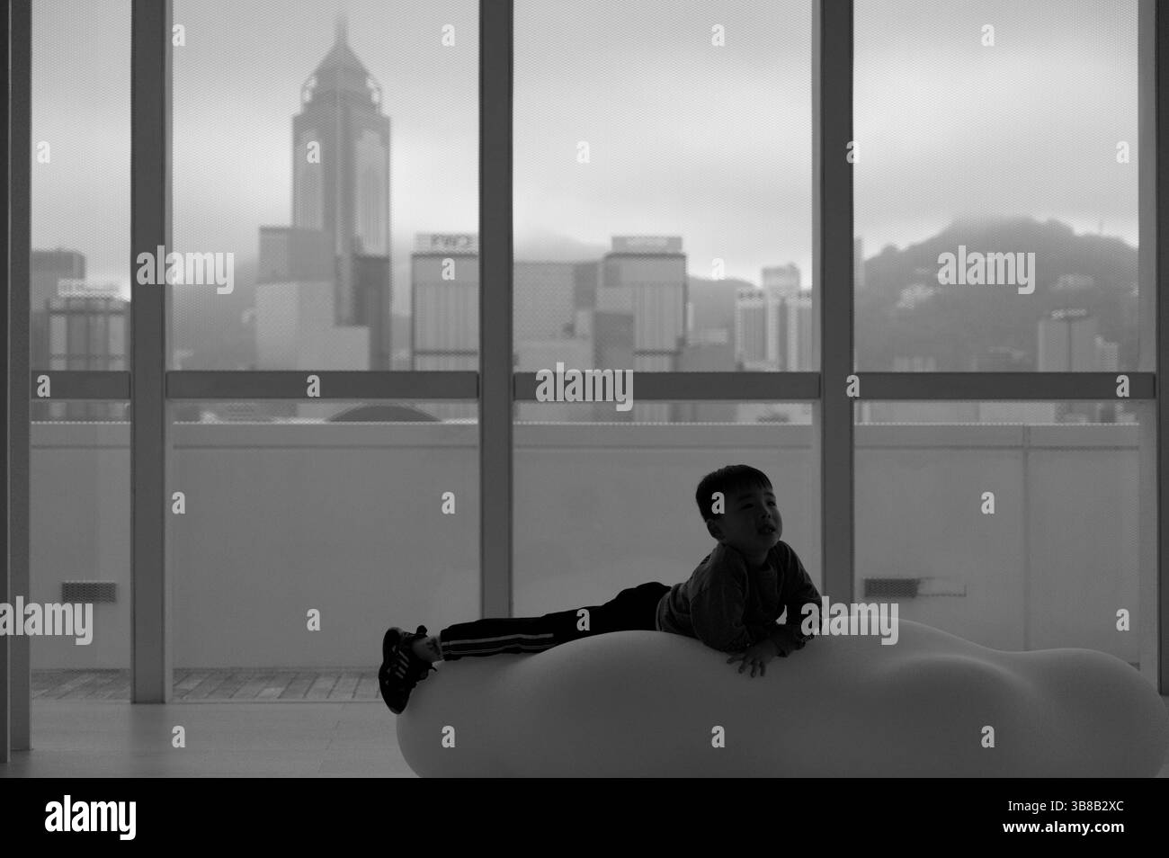 Ein Kind mit Silhouette zieht sich auf einem wolkenförmigen Sitz vor einer nebeligen Skyline der Stadt an der HKMoA Stockfoto