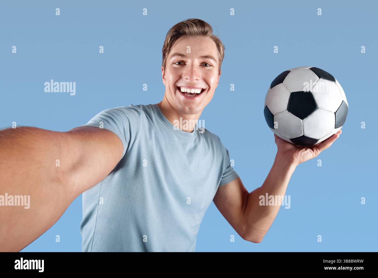 Lächelnder junger Mann mit Fußball nimmt Selfie auf seine Mobiltelefon über violettem Studiohintergrund Stockfoto
