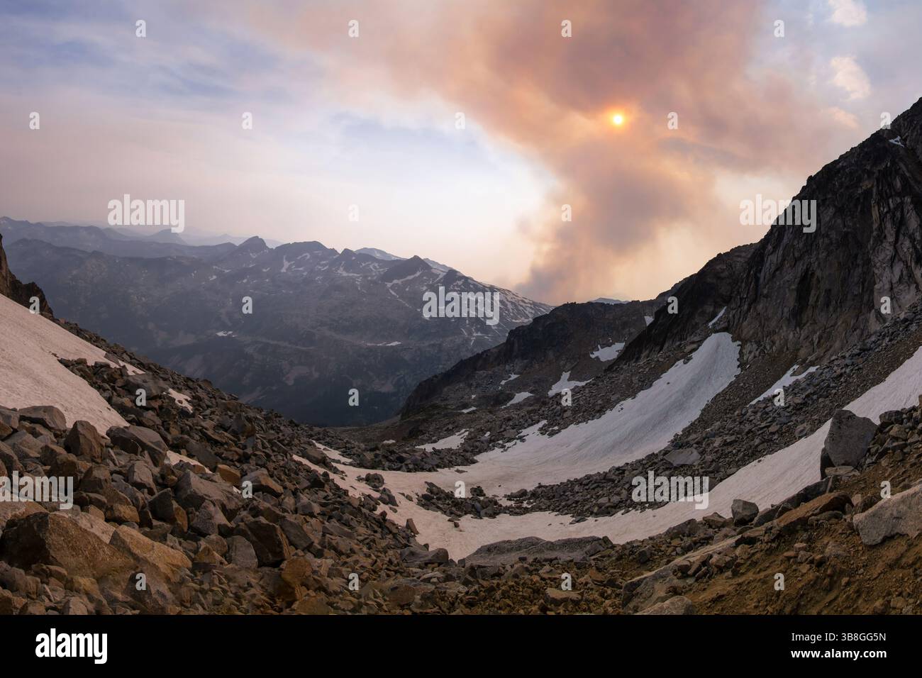 Eine atemberaubende Landschaft zeigt ein Tal voller Schnee und zerklüfteter Felsen, unter einem rauchigen Himmel, während die Sonne untergeht. Berge erheben sich in der Ferne, verbessern Stockfoto