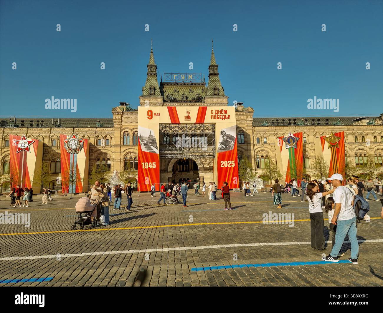 Menschen auf dem Roten Platz Moskau Russland, mit Dekorationen des Siegtages der Sowjetunion (2. Weltkrieg). Am 9. Mai 2025; dekoriertes KAUGUMMI im Hintergrund - Smartphone-aufgenommenes Stockfoto
