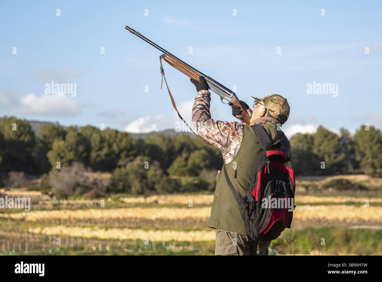 Hunter trägt Tarnkleidung und zielt auf das Feld auf dem Land während eines Tagesausflugs mit Bäumen und bewölktem Himmel im Hintergrund in Sant P Stockfoto