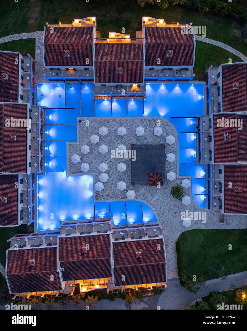 Blick aus der Vogelperspektive auf einen symmetrischen Hotelkomplex mit Pools, Sonnenschirmen und warm beleuchteten Gebäuden. Stockfoto