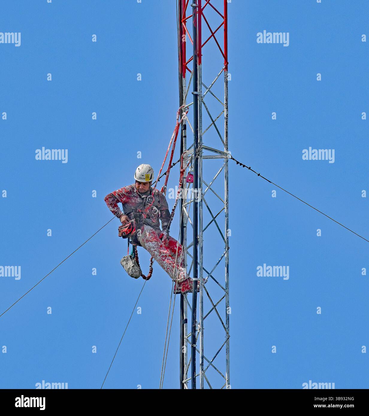 EMPORIA KANSAS, USA - 8. MAI 2025 der Turmtechniker, der mit Sicherheitsseilen in etwa 200 m Höhe an einem Funkturm eingehängt ist, verwendet einen Malhandschuh, um die Querträger weiß zu malen Stockfoto