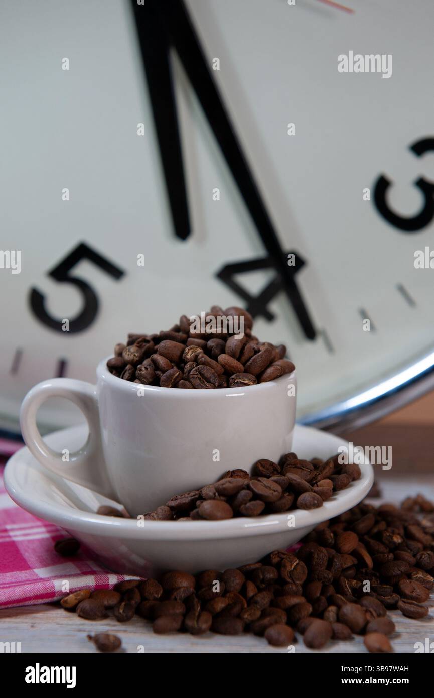 Eine kleine Kaffeetasse voller ganzer Kaffeebohnen und eine große industrielle Uhr Stockfoto