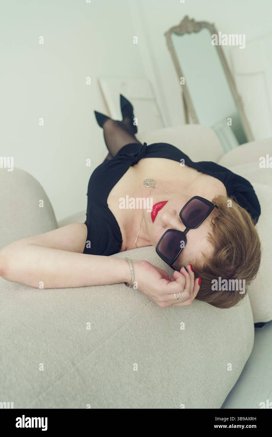 Eine erwachsene brünette Frau mit kurzen Haaren in einem schwarzen Kleid liegt auf dem Sofa und lehnt sich zurück Stockfoto