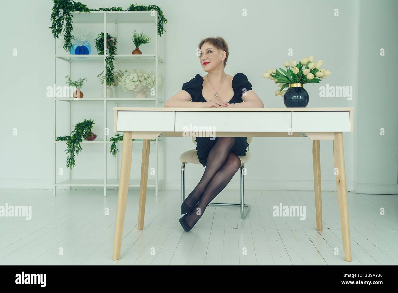 Eine Erwachsene, brünette Frau mit kurzen Haaren in einem schwarzen Kleid sitzt an einem Tisch und lehnt ihren Kopf auf ihre Hände Stockfoto