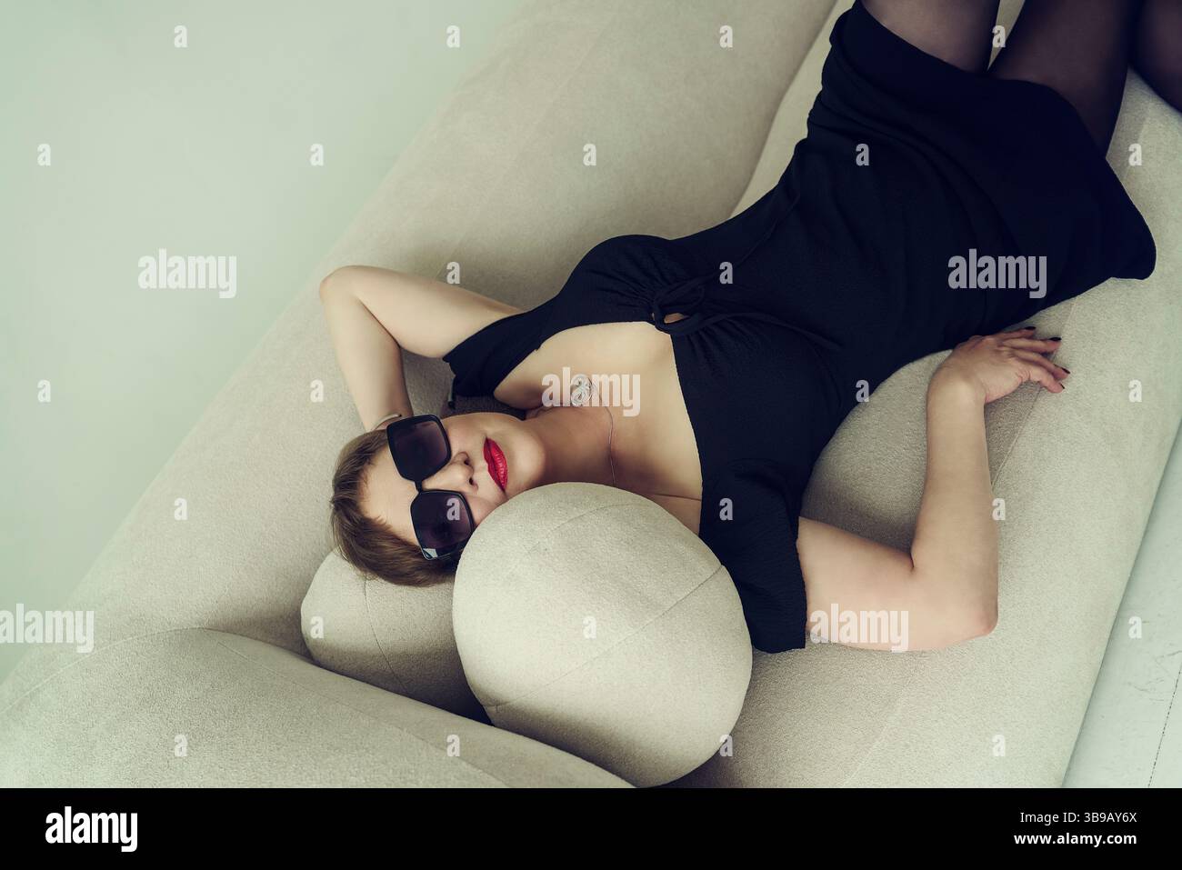 Eine erwachsene brünette Frau mit kurzen Haaren in einem schwarzen Kleid liegt auf dem Sofa und lehnt sich zurück Stockfoto