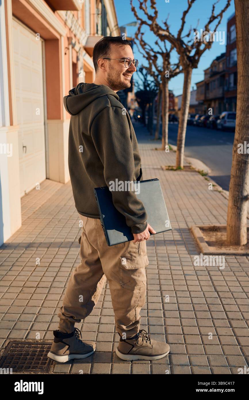 Lächelnder Mann, der auf dem Bürgersteig mit Laptop im Wohnbereich läuft Stockfoto