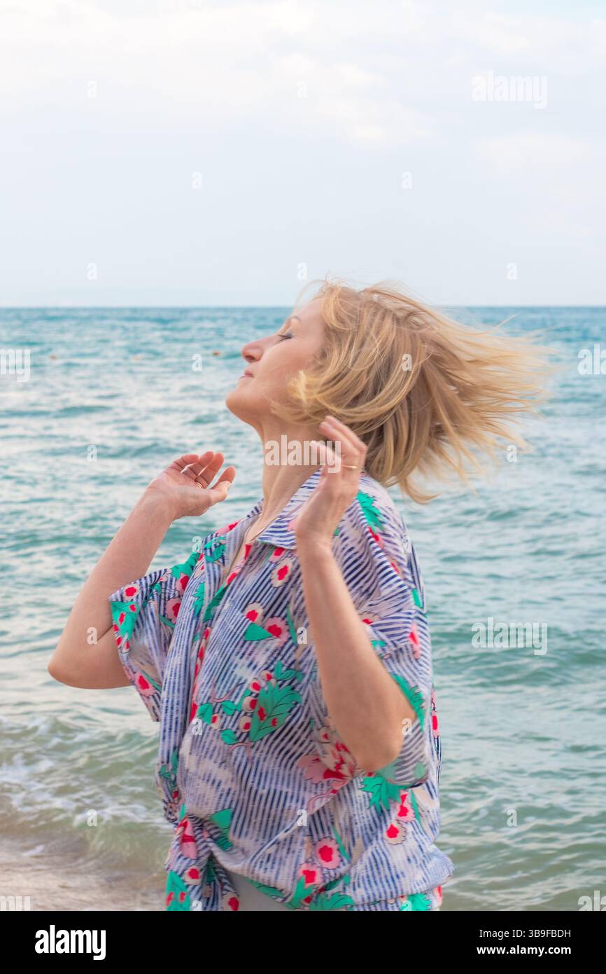 Frau mit kurzen blonden Haaren wirft Kopf zurück auf den Strand mit Ozean im Hintergrund. Unbeschwerter Sommerurlaub am Meer. Stockfoto