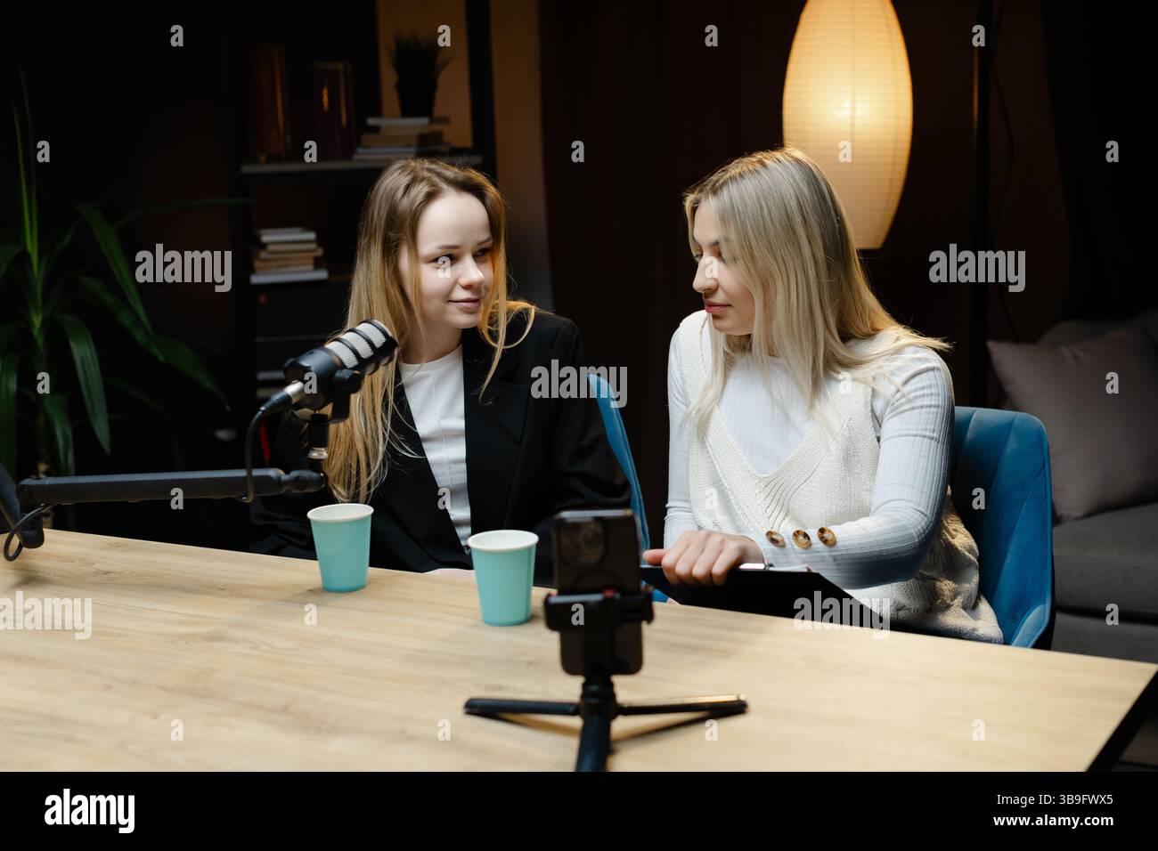 Zwei Podcasterinnen lächeln und nehmen eine Live-Show mit professionellem Mikrofon und Smartphone auf Stockfoto
